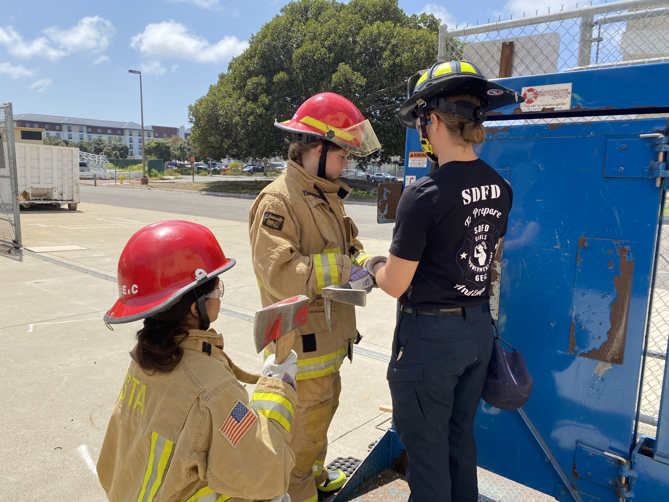 Picture of two teenage girls wearing yellow firefighter turnouts standing next to a female firefighter in a black shirt and black pants. All are wearing helmets and they are practicing forcible entry drills.